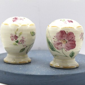 May include: A pair of white ceramic salt and pepper shakers with a floral design. The shakers have a gold trim and feature pink and green flowers.
