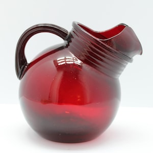 May include: A translucent ruby red glass pitcher with a spherical body, a curved handle, and a wide, angled spout. The neck of the pitcher features a series of horizontal ridges, and the glass has a smooth, polished finish throughout the piece.