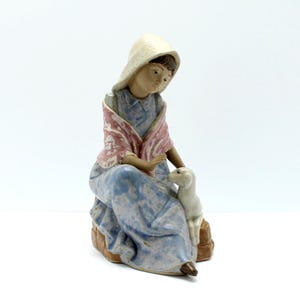 May include: A ceramic figurine of a seated figure with a white head covering and a patterned shawl. The figure is wearing a blue dress and is accompanied by a small white dog. The figurine is on a brown base.