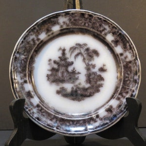 May include: A white porcelain plate with a black and white transferware design of a Chinese landscape. The plate has a scalloped edge and a black rim.