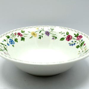 May include: A round white ceramic bowl featuring a delicate floral pattern along the inner rim. The design includes small green leaves and stems with colorful blossoms in shades of pink, yellow, blue, and purple, finished with a thin decorative border.