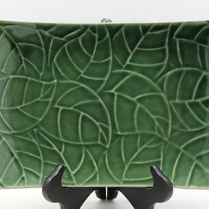 May include: A rectangular, green ceramic serving dish with a raised leaf pattern. The leaves are detailed with visible veins and are arranged in an overlapping design. The dish is displayed on a black stand against a white background.