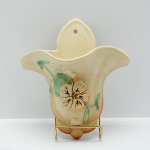 May include: A cream-colored ceramic wall pocket vase featuring a sculpted flower with green leaves and brown accents. The vase has a wide, flared opening and a single hole at the top for mounting. It is supported by a thin gold-toned metal stand.