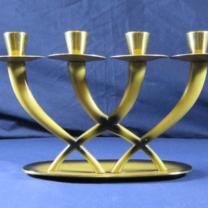 May include: A four-candle holder with a unique design. The candle holders are gold-colored and sit atop curved, light-yellow arms that intersect in the middle. The base is an oval shape, also light yellow with a gold trim.