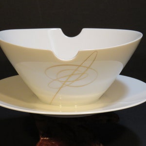 May include: A cream-colored gravy boat with a matching saucer. The boat has a unique, angular design with a spout and is decorated with gold abstract swirls. The set is made of porcelain.