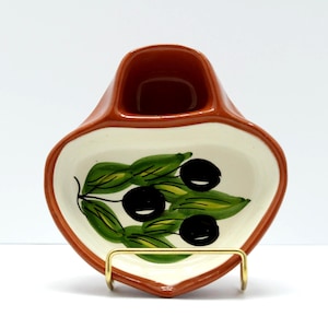 May include: Ceramic olive dish with a brown rim and a white interior. The dish features a painted design of green leaves and black olives. It has a small, rectangular compartment and is supported by a gold-colored metal stand.