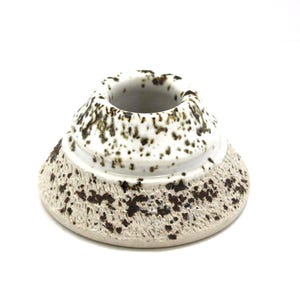 May include: A ceramic candle holder with a speckled design. The base is a light beige color with dark brown speckles, and the top is white with brown speckles. The candle holder has a wide base and a narrow top.
