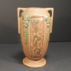 May include: A peach-colored ceramic vase with two handles and a pedestal base. The vase features a textured central panel and grape cluster designs in green and blue. The vase is a decorative item.