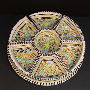 May include: A round, segmented serving dish with a central bowl and six surrounding compartments. The dish is white with a yellow border and intricate geometric patterns in red, blue, and green. The rim has a black dotted design.