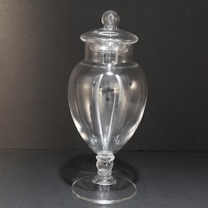May include: A clear glass apothecary jar with a lid. The jar has a bulbous body, a slender stem, and a round base. The lid is topped with a small, round knob. The jar is likely used for storing items.