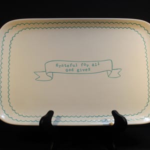 May include: Cream-colored rectangular tray with a scalloped teal border and the text "grateful for all god gives" in a banner. The tray is displayed on a black stand against a black background.
