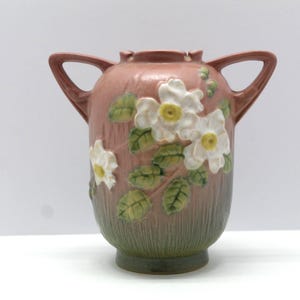 May include: A ceramic vase with a pinkish-brown body, adorned with white floral relief and green leaves. It features two arched handles and a textured, green base. The vase is approximately 10 inches tall.