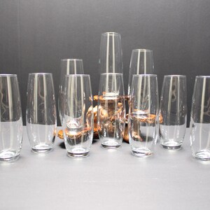 May include: A collection of clear, stemless drinking glasses. The glasses are cylindrical with a slightly rounded shape. Some glasses contain liquid. The glasses are arranged on a gray surface with a dark background.