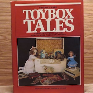 May include: A hardcover book titled Toybox Tales by Virginia S. Carr with a red cover. The front features a photograph of a white toy chest filled with vintage dolls, a stuffed animal, and books, surrounded by a doll, a toy piano, and other toys.