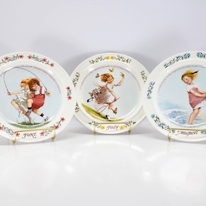 May include: Three decorative plates featuring illustrations of children at play. Each plate has a white background with a floral border and a central image depicting a different activity. The plates are labeled "June", "July", and "August".