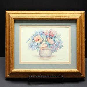 May include: A framed watercolor painting featuring a bouquet of soft pink and blue flowers in a light-colored vase. The artwork is encased in a light brown wooden frame with a light blue matting and a cream-colored inner border.