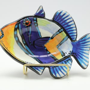 May include: A ceramic fish-shaped dish with vibrant colors. The dish features a design with orange, yellow, blue, and purple hues. It is supported by a gold-colored metal stand. The dish has the text "Humuhumunukunukuapua'a" and "Hawaii 2004".