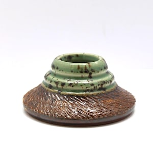 May include: A handcrafted ceramic vase with a unique design. The vase features a light green top with dark speckles and a textured, brown base. The vase has a circular opening and a wide, flat base.