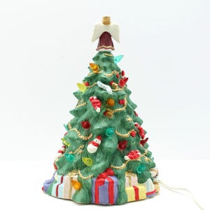 May include: A ceramic Christmas tree with green branches, adorned with colorful lights, ornaments, and a gold garland. A white angel tops the tree. Presents are at the base. A festive holiday decoration.