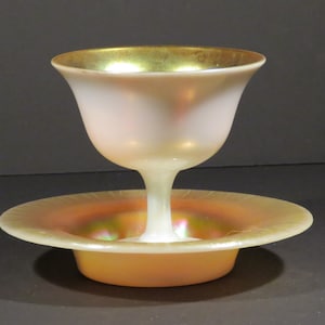 May include: An iridescent glass compote with a gold rim. The compote features a flared bowl atop a slender stem, resting on a matching saucer. The glass has a pearlescent sheen, reflecting colors of orange, pink, and green.