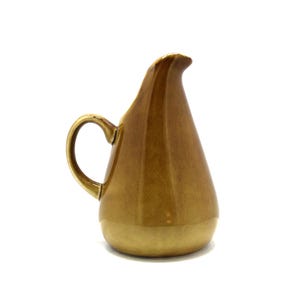 May include: A vintage, teardrop-shaped pitcher with a curved handle and a spout. The pitcher is a warm, golden-brown color with a lighter base. The ceramic pitcher is likely used for serving beverages.