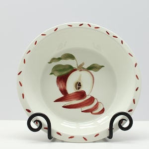 May include: A white ceramic pie plate with a hand-painted apple design. The apple is red with green leaves and sliced to show the inside. The rim has a dotted red pattern. The plate is displayed on a black metal stand.