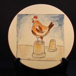 May include: A ceramic plate with a hand-painted design of a rooster standing on a bucket. The rooster is brown and white with a red comb and wattle. The background is a light blue watercolor wash.