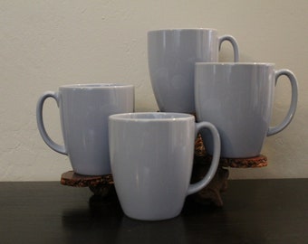 May include: Four light blue ceramic mugs with handles, arranged on a wooden coaster.