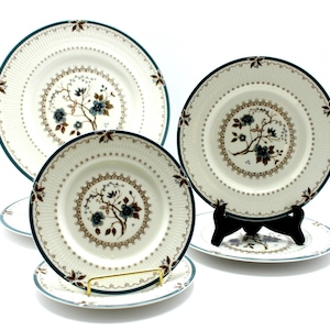 May include: A collection of cream-colored ceramic plates with a floral design. The plates have a dark teal border and a detailed floral pattern in the center. The plates are displayed on stands.