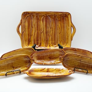 May include: A set of ceramic serving dishes in the shape of corn on the cob. The set includes a large rectangular platter with handles and six individual corn-shaped dishes. The dishes are glazed in shades of brown and yellow.