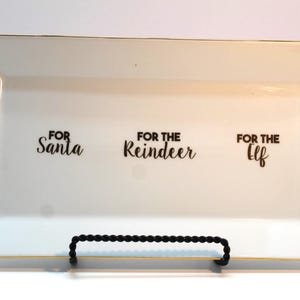 May include: A rectangular white serving tray with gold trim and black text that reads "FOR Santa", "FOR THE Reindeer", and "FOR THE Elf". The tray is supported by a black metal stand.