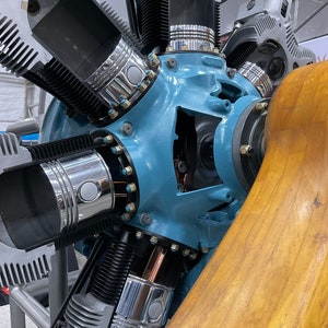 May include: A close-up of a vintage aircraft engine with a blue painted casing and silver pistons. The engine is connected to a wooden propeller.