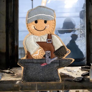 May include: A felt ornament depicting a blacksmith, with a smiling face, wearing a blue cap, brown overalls, and a black glove. The blacksmith is working on an anvil with a hammer. The ornament is tan, brown, grey, and white.