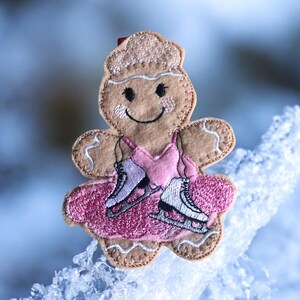 May include: A gingerbread person wearing a pink dress and ice skates. The gingerbread person is smiling and has a brown face. The gingerbread person is on a white background.
