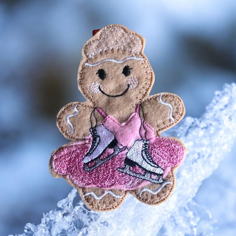May include: A gingerbread person wearing a pink dress and ice skates. The gingerbread person is smiling and has a brown face. The gingerbread person is on a white background.