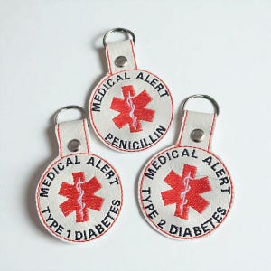 May include: Three white medical alert keyrings with red stitching and a silver key ring. Each keyring features a red medical alert symbol and black text indicating a medical condition: Penicillin, Type 1 Diabetes, and Type 2 Diabetes.
