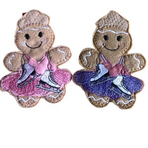 May include: Two gingerbread figure ornaments, one with a pink dress and the other with a purple dress, both wearing ice skates.