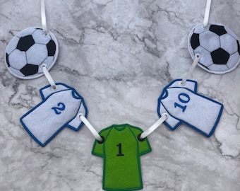 Puede incluir: Una guirnalda de cinta blanca con recortes de fieltro de balón de fútbol y camiseta. Las camisetas tienen los números 1, 2 y 10.