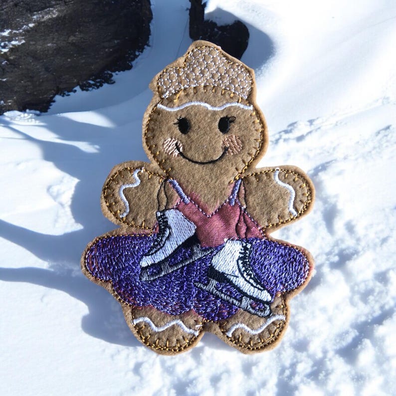May include: A gingerbread figure wearing a purple dress and ice skates. The figure has a smiling face and a brown felt body.