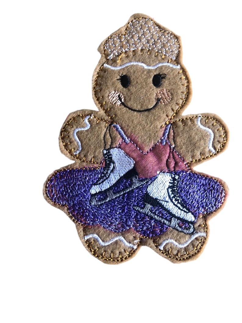 May include: A gingerbread person wearing a purple dress and ice skates. The gingerbread person has a smiling face and a white icing hat.