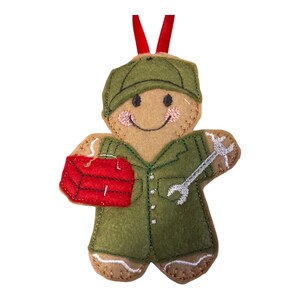 May include: A gingerbread person ornament with a green uniform, a red toolbox, and a wrench. The gingerbread person is smiling and wearing a green hat.