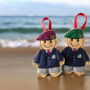 May include: Two felt ornaments shaped like people wearing military uniforms. One ornament has a burgundy beret and the other has a green beret. Both ornaments have a smiling face and are decorated with gold buttons.