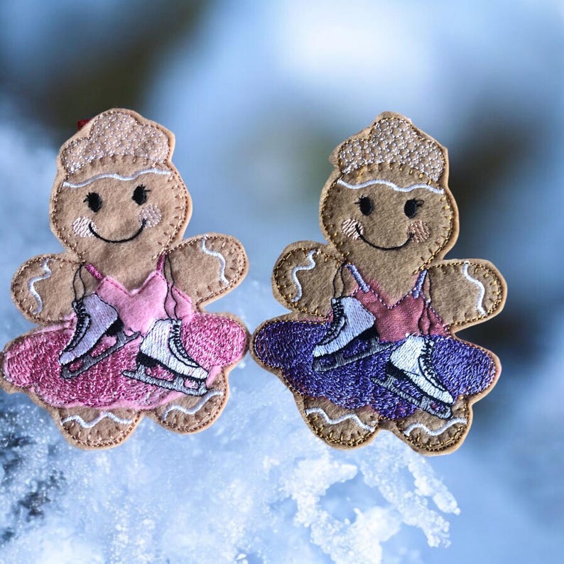May include: Two gingerbread ornaments decorated with embroidered details. One ornament is wearing a pink dress and the other is wearing a purple dress. Both ornaments are holding ice skates.