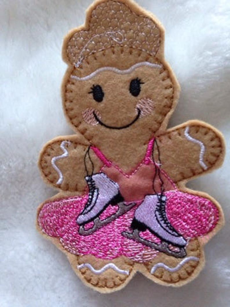 May include: A felt gingerbread person ornament wearing a pink dress and ice skates. The ornament is decorated with white stitching.