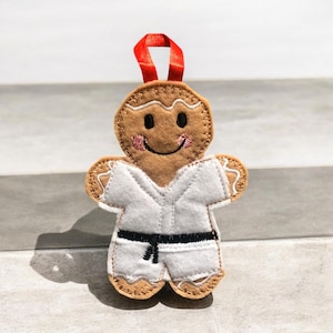 May include: A gingerbread person ornament wearing a white karate uniform with a black belt. The ornament has a smiling face and a red ribbon for hanging.
