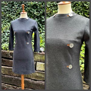 May include: A charcoal gray dress with a high neckline and three-quarter sleeves. The dress features large, round buttons in a contrasting color. The dress is displayed on a mannequin against a backdrop of greenery.