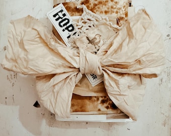 May include: A stack of aged, tea-stained paper ephemera tied with a large, cream-colored fabric bow. The bundle includes a white tag with the word HOPE printed in black, a small piece of white crochet lace, and a paper featuring a bird illustration.