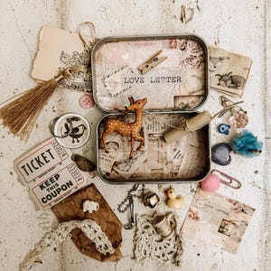 May include: A collection of vintage-style ephemera, including a metal tin labeled "LOVE LETTER", a small deer figurine, tickets, lace, and a yellow rubber duck. The items are arranged on a distressed white wooden surface.