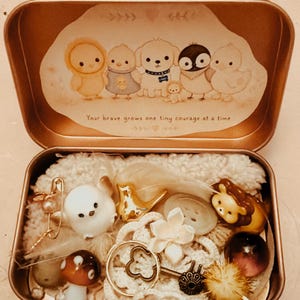May include: A rectangular metal tin containing a collection of small trinkets including a white bird figurine, a gold bird, a mushroom charm, a lion figure, and a key. The lid features an illustration of cartoon animals and the text: Your brave grows one tiny courage at a time.