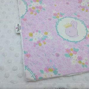 Puede incluir: Una manta de beb&eacute; blanca con un estampado floral rosa en un lado. El estampado floral presenta una corona y un conejito. La manta tiene una etiqueta que dice "Starline Designer".